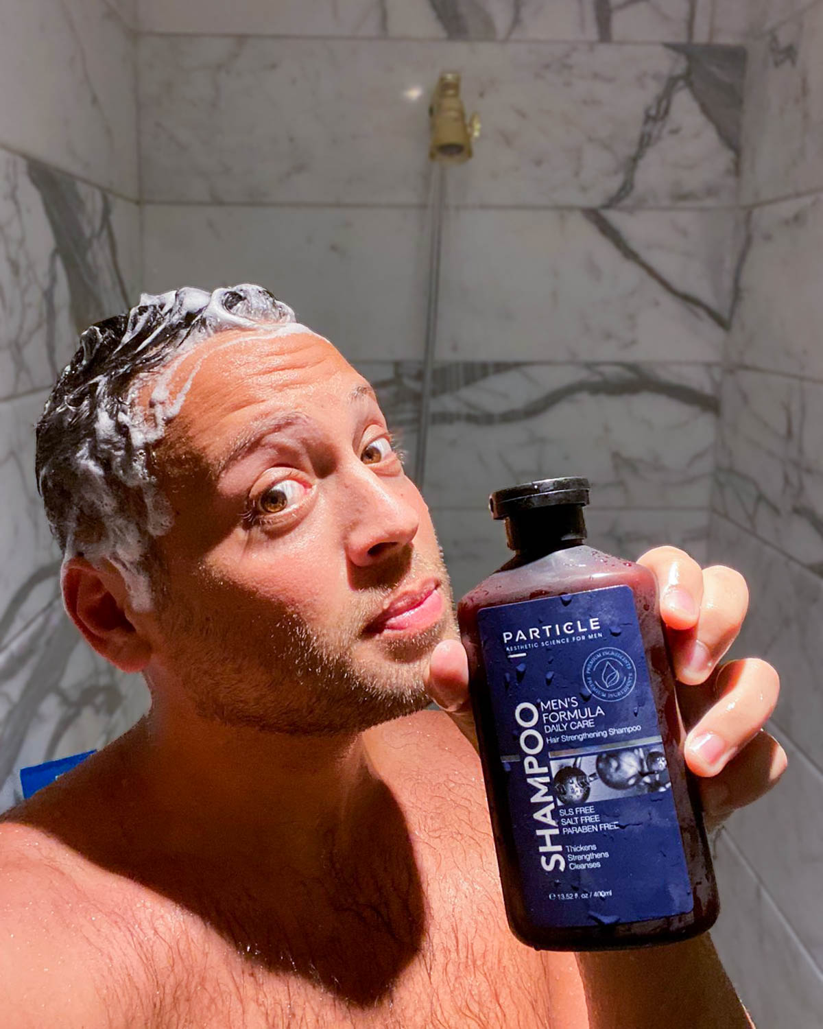 Man with soapy hair holding Particle shampoo bottle in the bathroom.