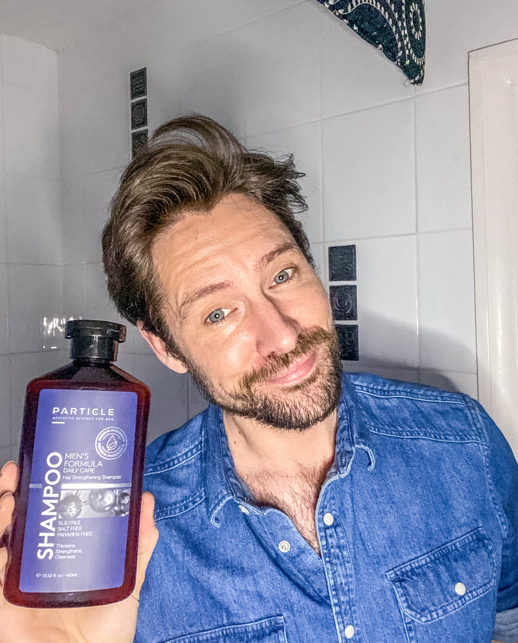 A man in blue shirt, smiling, holding a bottle of Particle Men's Formula Hair Strengthening shampoo.