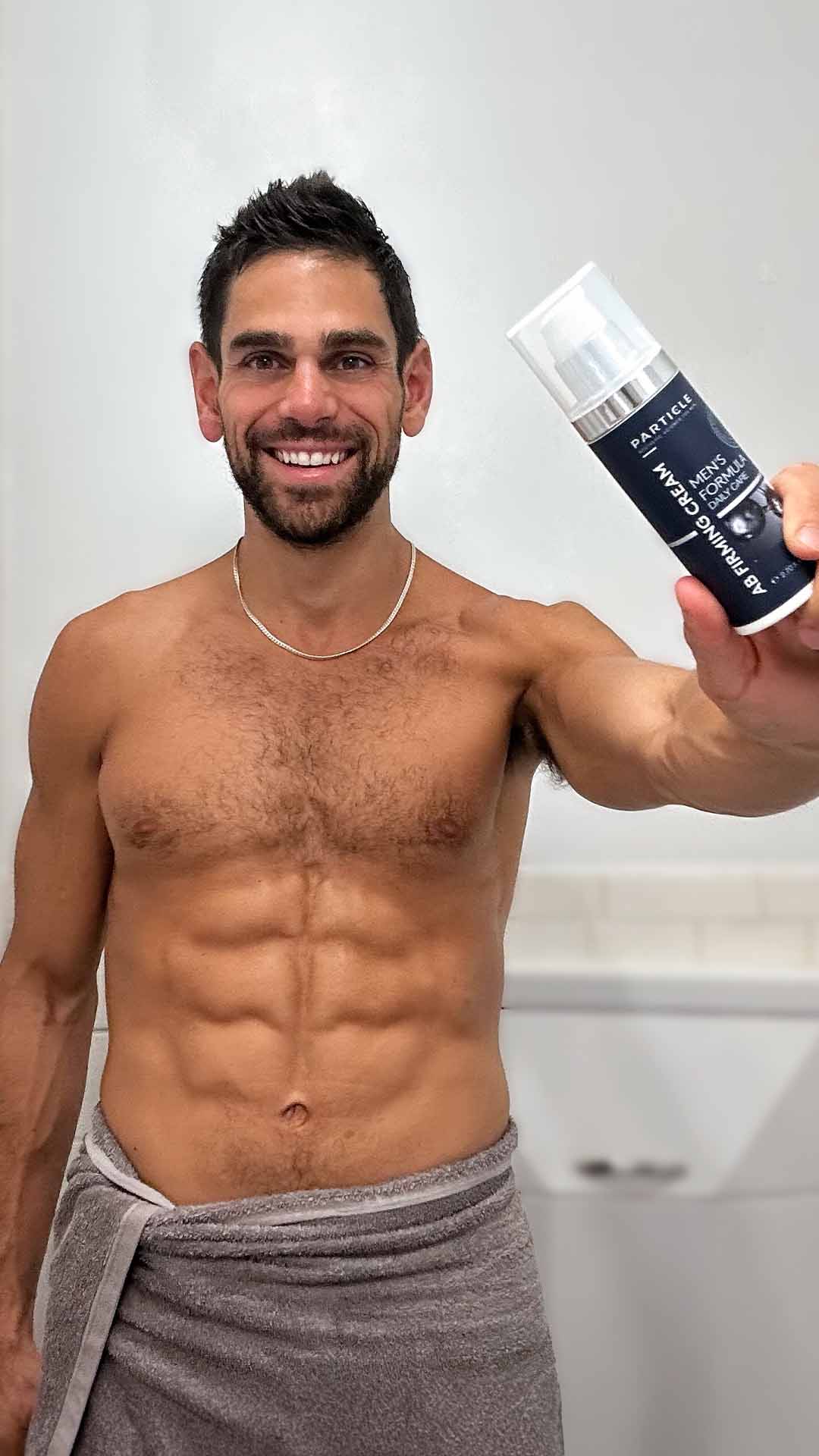 Man with defined abdominal muscles, smiling, holding Particle AB Firming Cream bottle.