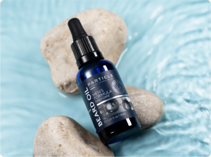 A blue bottle labeled Particle Beard Oil with a black dropper cap on light-colored rocks.